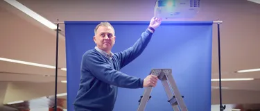 Dieses Bild zeigt Jörg Lohmann, der auf einer Leiter steht und eine Hand an einen Beamer hält.