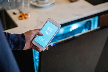 Smarte Geschirrspüler. Dieses Bild zeigt den Ausschnitt einer Person, die ihr Handy in der Hand hält. Im Hintergrund ist ein Geschirrspüler zu sehen.