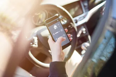 Smarte Heizung. Dieses Bild zeigt eine einen Ausschnitt einer Person, die im Auto hinterm Steuer sitzt und an ihrem Handy ist.