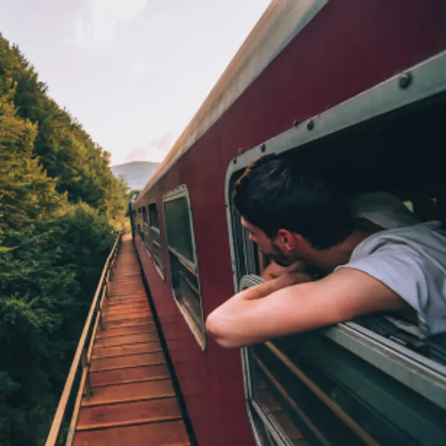 Mann schaut aus dem Zugfenster Wer das öffentliche Verkehrsnetz für einen Urlaub nutzt, tut dem Klima etwas Gutes. Zu sehen ist ein Mann, der aus dem Fenster eines fahrenden Zuges schaut.
