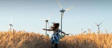 Die Zukunft unserer Energie. Das Bild zeigt ein Mädchen, das durch ein Getreidefeld, auf dem Windkraftanlagen stehen, läuft. 