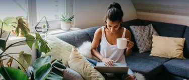 Klimafreundlich kühlen. Dieses Bild zeigt eine junge Frau auf dem Sofa. In der Hand hält sie eine Tasse und auf dem Schoß einen Laptop. Um sie herum stehen Pflanzen.