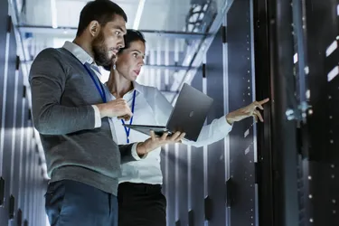 Eine Frau und ein Mann der ein Laptop in den Händen hält stehen in einem Rechenzentrum vor einem Server und kontrollieren dessen Werte