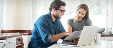 Ein Ehepaar sitzt an einem Tisch vor dem Laptop und schaut sich Papiere an.