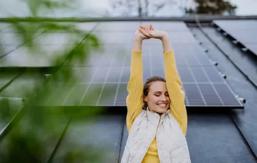 Eine junge Frau steht vor einer mit PV-Modulen bestückten Hausfassade und streckt ihre Arme zufrieden in die Luft 
