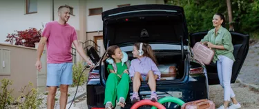 Eine Familie bereitet sich auf den Urlaub vor: Der Vater tankt das E-Auto, die Mutter verstaut gepäck im E-Auto und zwei Töchter sitzen spielend im Kofferraum.