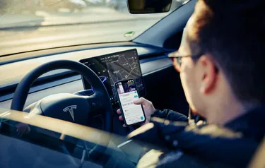 Ein in seinem Auto am Steuer sitzender Mann nutzt an seinem Handy die &Charge-App