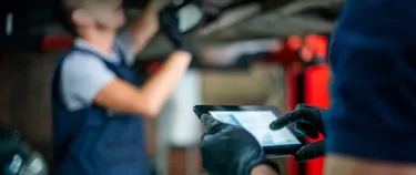 Das Bild zeigt einen Mechaniker in der Werkstatt unter einem Auto. Daneben steht ein weiterer Mechaniker und trägt auf einem Tablet den Batterie Pass ein. 