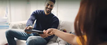 Eine Frau reicht einem Mann ein Tablet, während beide Personen auf einem Sofa sitzen.