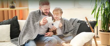 Ein Vater und seine Tochter sitzen mit Teetassen in der Hand unter einer Decke auf dem Sofa und lächeln.