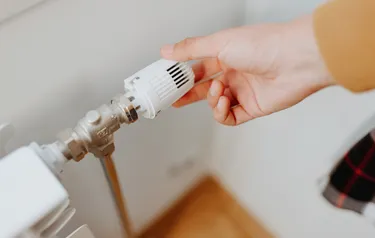 Eine Person regelt das Heizungsthermostat an einem Heizkörper, welches auf Stufe 0 eingestellt ist