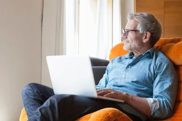 Ein älterer Mann sitzt mit dem Laptop auf dem Schoß in einem Sessel und blickt lächelnd aus dem Fenster