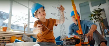 Ein Junge und sein Opa spielen mit einem Spielzeug-Kran und einem Spielzeug-Strommast. Beide tragen Sicherheitshelme.