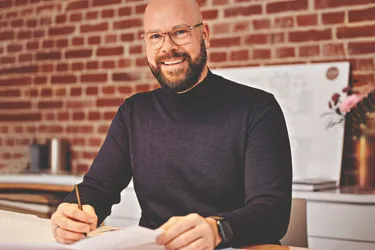 Das Bild zeigt den Architekten Dennis Fleer, der lächelnd an einem Tisch sitzt, auf welchem ein Bauplan ausgebreitet ist.