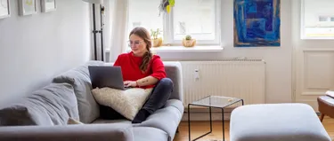 Eine Frau sitzt auf ihrem Sofa und schaut auf ein Laptop.