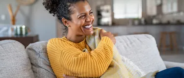 Eine Frau sitzt lächelnd mit einer Decke auf dem Sofa.