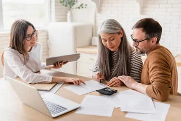 Eine Frau mit Unterlagen in der Hand berät eine Frau und einen Mann die ihr gegenüber am Tisch sitzen und sich gemeinsam mit ihr über Unterlagen beugen