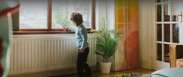Header: Ein kleiner Junge steht in einer Wohnung vor einer Heizung unter einem Fenster und spielt mit einem Dino auf der Heizung.