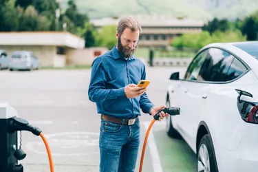Ein Mann nutzt neben seinem an einer Ladesäule tankenden E-Auto stehend mit seinem Mobiltelefon die &Charge-App