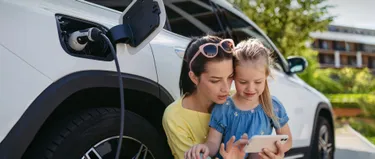 Eine Mutter zeigt ihrer Tochter etwas auf ihrem Handy, während im Hintergrund ein E-Auto lädt.