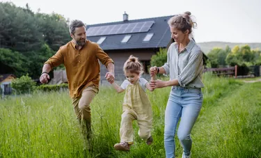 Ein Mann und eine Frau laufen durch den Garten eines Einfamilienhauses und wirbeln dabei ihre Tochter an den Händen durch die Luft