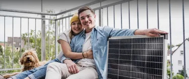 Ein junges Pärchen sitzt mit seinem Hund und einer Mini-Solaranlage auf dem Balkon.