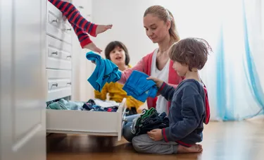 Nachhaltig wohnen bedeutet auch, regelmäßig den Kleiderschrank im Kinderzimmer auszumisten. Das Bild zeigt eine Mutter mit ihren beiden Kindern, während sie eine Kleiderkommode aufräumen.