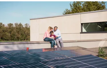 Eine Frau ein Mann und ein Kind sitzen auf dem Dach ihres Hauses auf dessen Flachdach eine PV-Anlage montiert wurde
