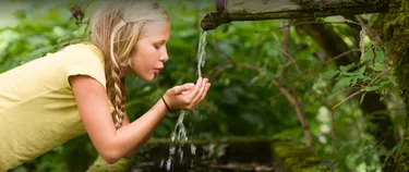 Ein Mädchen fängt Wasser, welches mitten im Wald aus einem kleinen Rohr fließt, mit ihren Händen auf. 