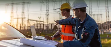 Zwei Männer mit Arbeitskleidung und Schutzhelm sehen sich Pläne auf Papier und einem Laptop an und zeigen auf Stromleitungen im Hintergrund.
