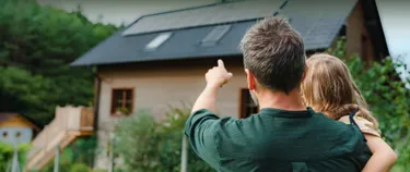 Ein Vater hat sein Kind auf dem Arm und zeigt auf ein Haus, dessen Dach mit einer Solaranlage ausgestattet ist.