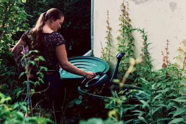 Eine Frau steht am Haus im Garten und gefüllt eine Gießkanne in einer Regentonne, die an die Dachrinne angeschlossen ist.