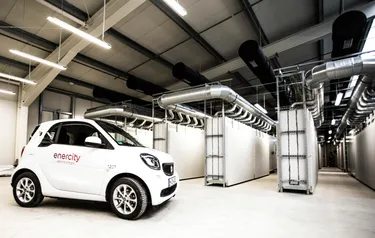 Ein mit dem enercity-Logo versehenes weißes E-Auto der Marke Smart steht vor einer Reihe von Batterien im Inneren des Batteriespeichers Herrenhausen