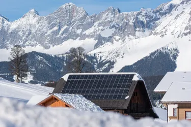 Blick auf eine winterlich verschneite Berglandschaft in deren Vordergrund ein Haus steht dessen Dach mit einer Solaranlage bestückt ist