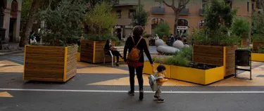 Eine Mutter und ihr Sohn laufen durch einen mit Grünflächen und Sitzgelegenheiten ausgestatteten Platz.