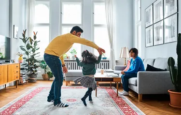 In einem Wohnzimmer tanzt ein Vater mit seiner kleinen Tochter auf einem Teppich während die Mutter vom Sofa aus zuschaut