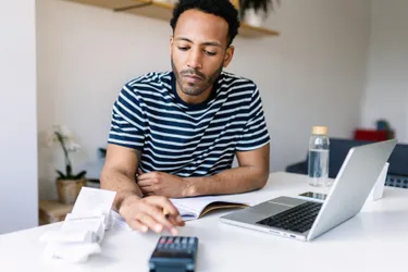 Ein Mann tippt am Schreibtisch neben einem Laptop sitzend auf einem Taschenrechner