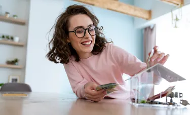 Eine Frau steckt Geldscheine in eine Spardose aus Plexiglas, die die Form eines Hauses hat