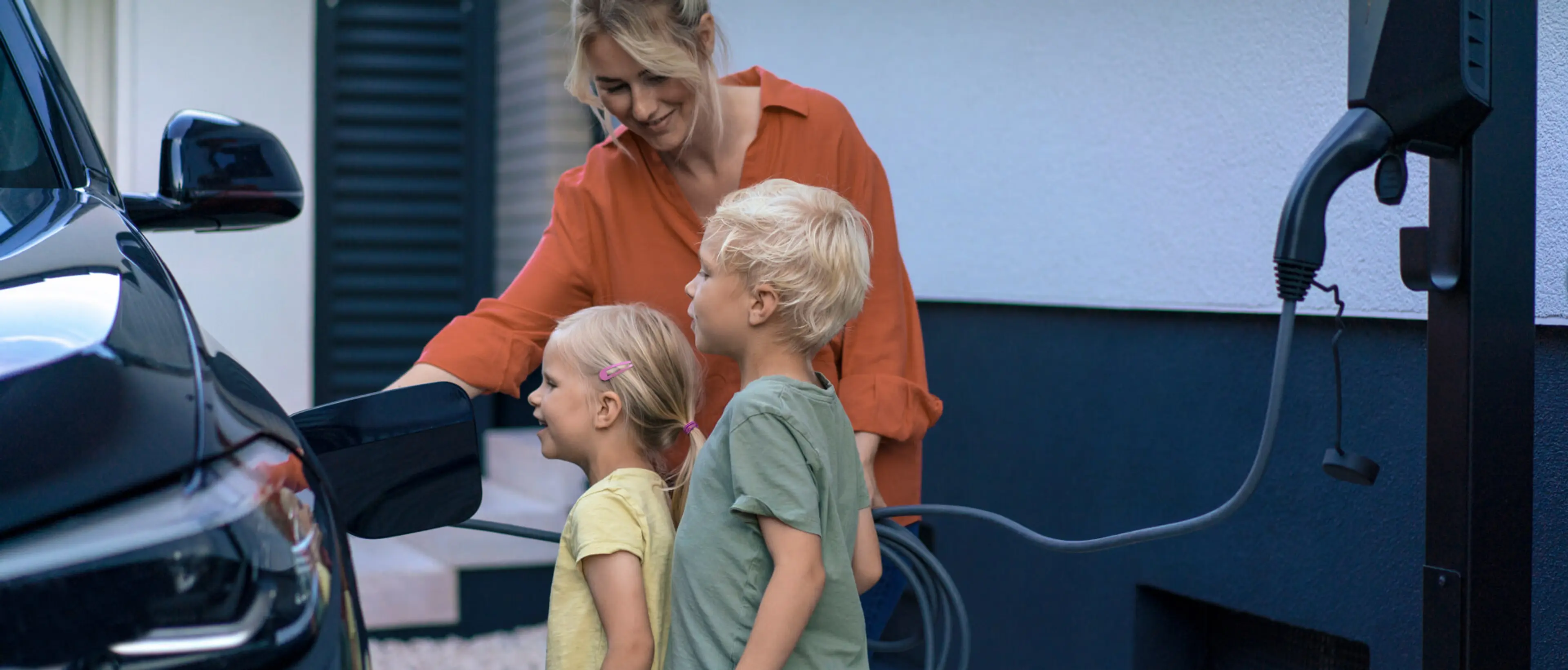 Wallbox Eine Mutter lädt ihr E-Auto, während ihre beiden Kinder, ein Junge und ein Mädchen, zuschauen.