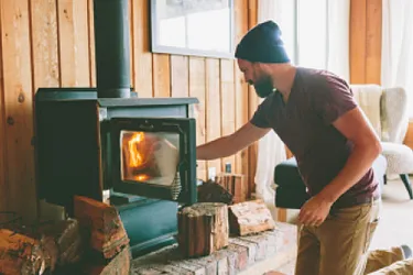 Dieses Bild zeigt einen Mann, der Holz in einen brennenden Kaminofen legt.